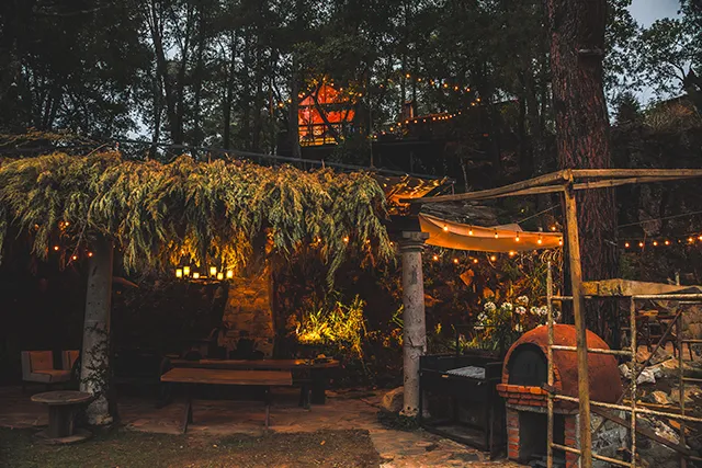 Una terraza rústica con horno de barro, iluminada por luces colgantes, bajo una cabaña en el árbol visible entre los pinos al anochecer.