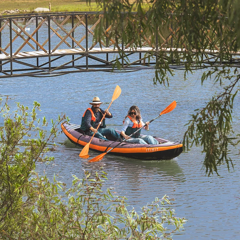 En la imagen, dos personas en chalecos salvavidas reman un kayak inflable naranja y negro. De fondo, hay un puente de madera y follaje.