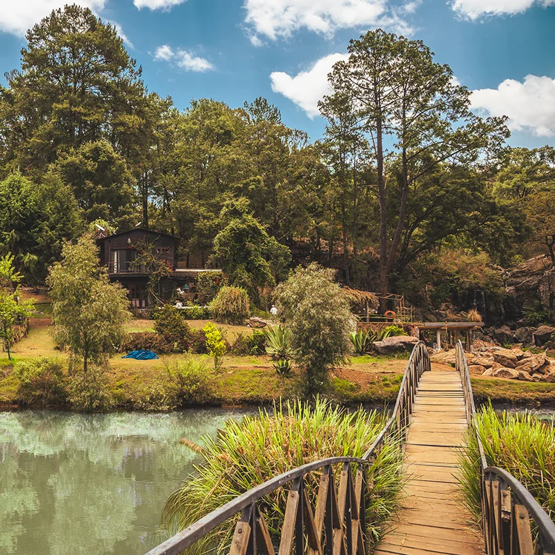 Un puente de madera cruza un cuerpo de agua hacia una casa con balcón, rodeada de árboles y bosque bajo un cielo nublado.