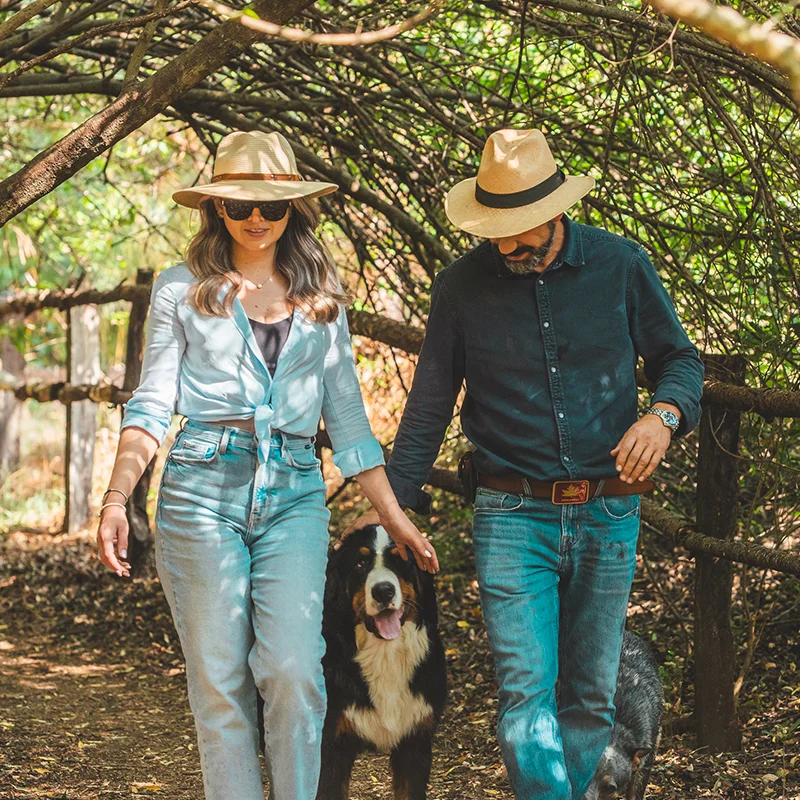 La imagen muestra a un hombre y una mujer caminando por un sendero arbolado con un perro bernés de la montaña. Ambos llevan sombreros de paja y ropa casual; la mujer usa lentes de sol.