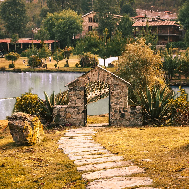 La imagen muestra un camino de losas de piedra que lleva a una estructura de arco de piedra rústica. Detrás de esta, hay un lago tranquilo y al fondo, se ven casas entre árboles, bajo una luz cálida.