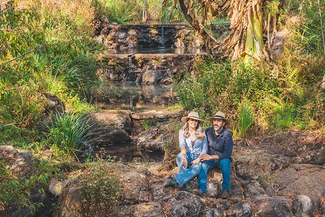Una pareja con sombreros se sienta junto a un arroyo en un entorno natural frondoso con cascadas.